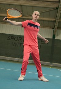 Korálový sportovní set obsahuje tričko s krátkým rukávem a námořnickým pruhem, doplněné o odpovídající kalhoty a bílé tenisky na tenisovém kurtu.