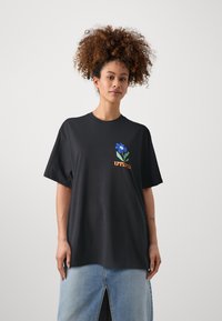 Camiseta de algodón negra con mangas cortas, que presenta un diseño de flor azul y el texto "UTOPÍA" en naranja. Textura suave y corte relajado.