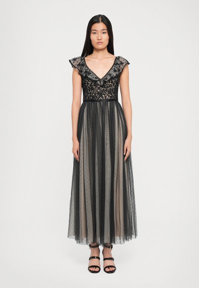 Frau in einem ärmellosen schwarzen Spitzenkleid mit Rüschenausschnitt und transparentem gepunktetem Overlay, vor einem schlichten Hintergrund stehend.