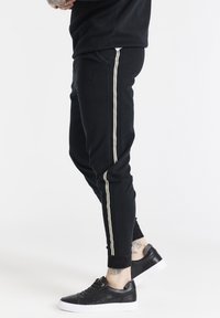 Svarta träningsbyxor med krämfärgade sidostråk, med en broderad logotyp på låret, mjukt tyg och ribbade ärmslut. Bar med svarta sneakers.