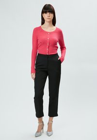 Rosa gerippter Cardigan mit Knöpfen und Verzierungen, kombiniert mit schwarzen maßgeschneiderten Hosen und silbernen High-Heels. Alles mit glatten Oberflächen.