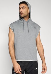 Ανοιχτό γκρι φούτερ χωρίς μανίκια από βαμβάκι με κουκούλα και κορδόνι, διακοσμημένο με μικρό λογότυπο στο στήθος και αντίθετη διακόσμηση στο τελείωμα.