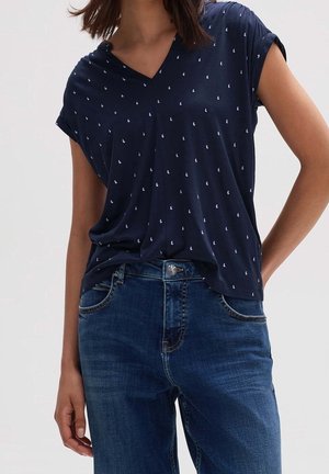 Frau trägt ein marineblaues V-Ausschnitt-Shirt mit kleinen weißen Mustern, das in eine blaue Jeans aus Denim gesteckt ist, vor einem schlichten Hintergrund.