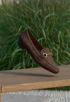 Mocassini in suede marrone con un design elegante, caratterizzati da un dettaglio in metallo dorato e un tacco basso, su uno sfondo verde sfocato.