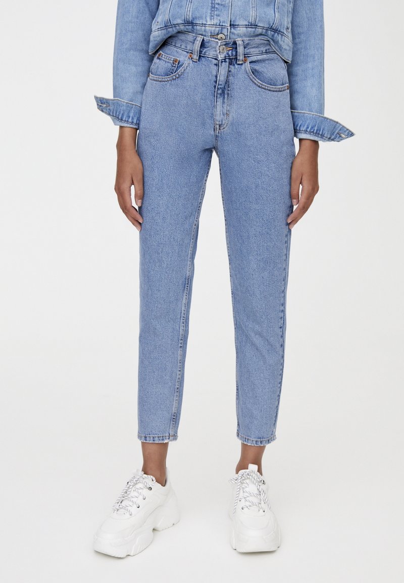 Personne portant un jean slim taille haute bleu clair et une veste en jean courte avec des baskets blanches épaisses sur un fond uni.