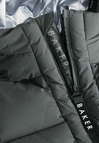 Graue gepolsterte Jacke mit hohem Kragen und seitlichem Reißverschluss, mit schwarzem Hardware und Branding entlang des Reißverschlussbands. Glatte Textur.