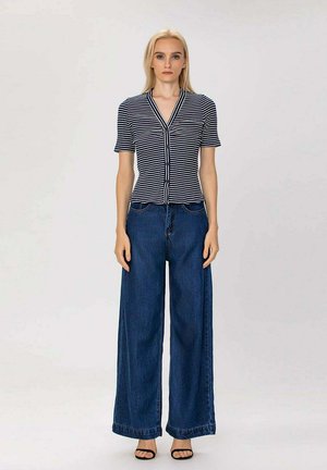 Femme debout portant un haut à rayures noires et blanches à manches courtes et boutonné, un jean bleu à jambes larges, et des talons ouverts noirs sur un fond uni.