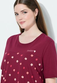 Frau mit langen braunen Haaren, die ein kastanienbraunes Shirt mit mehrfarbigem Blumenmuster und dem Schriftzug "SWEET" mit einem Herz auf der Brust trägt.