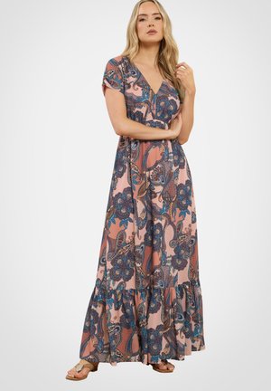 Vrouw in een roze maxi-jurk met blauwe paisley bloemenpatronen, korte mouwen en een gerimpelde zoom, staand met één hand die haar haar aanraakt.