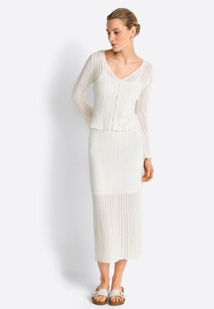 Woman with blonde hair in a white buttoned knit cardigan and matching long knit skirt, standing with hands behind back, wearing white sandals.