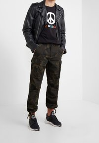 Zwarte leren motorjack over een zwart T-shirt met een vredesteken, gecombineerd met camouflage cargobroeken en zwarte sportschoenen.