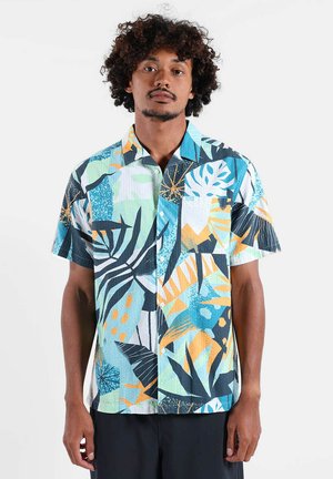 Homme aux cheveux bouclés portant une chemise à manches courtes avec des motifs abstraits de feuilles tropicales en tons bleus, orange et blancs.