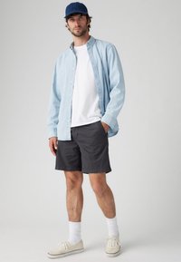 Cămașă albastră deschisă cu nasturi peste un tricou alb, pantaloni scurți gri, ciorapi albi și pantofi sport crem. O șapcă albastră adaugă contrast ținutei.