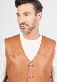 Bruine leren vest met een V-halsontwerp, voorzien van twee roze knopen en opvallende schoudernaden. Draag het over een lichtgekleurde gebreide shirt.