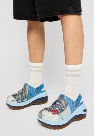 Pieds portant des chaussettes blanches et des sabots à plateforme bleus et gris avec des boutons décoratifs, assortis à un pantalon noir court.
