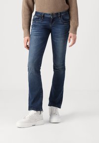 Personne portant un jean bleu foncé ajusté, un pull en laine beige et des baskets blanches, debout devant un fond clair uni.