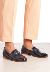 Dark navy loafers made of patterned patent leather with a buckle accent, paired with peach-colored cropped pants. Rubber sole visible.