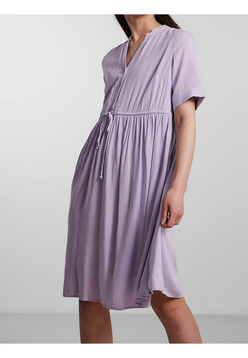 Femme portant une robe lavande ample, longueur au genou, avec des manches courtes et un lien à la taille, debout devant un fond uni gris clair.