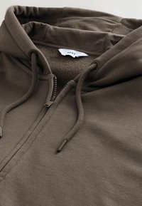 Sudadera con cremallera de color marrón hecha de una tela suave, con capucha ajustable y cremallera en la parte delantera. El interior tiene una textura cepillada. Etiqueta visible.