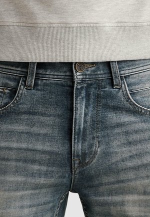 Detailný záber na stred tela v opraných modrých džínsoch s gombíkom a očkami na opasok, v kombinácii so svetlosivým rebrovaným ukončením svetra.