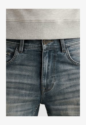 Detailný záber na stred tela v opraných modrých džínsoch s gombíkom a očkami na opasok, v kombinácii so svetlosivým rebrovaným ukončením svetra.