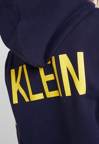 Mörkblå hoodie med huva, som visar stora gula versaler som stavar "KLEIN" på baksidan.