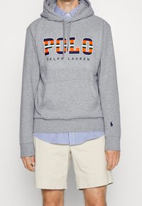Grå huvtröja med "POLO RALPH LAUREN"-grafik. Har en känguruficka, ribbade ärmslut och är lagd över en ljusblå randig skjorta och beiga shorts.