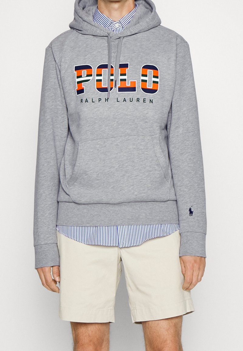 Grå huvtröja med "POLO RALPH LAUREN"-grafik. Har en känguruficka, ribbade ärmslut och är lagd över en ljusblå randig skjorta och beiga shorts.