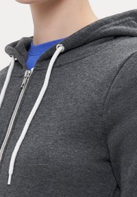 Sudadera gris con cremallera y capucha con cordones, con una tela texturizada y cordones blancos, usada sobre una camiseta azul.