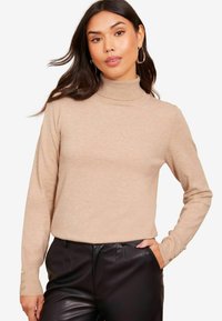 Beige rolák vyrobený z měkkého pleteného materiálu, s dlouhými rukávy a knoflíkovými detaily na manžetách, v kombinaci s černými koženými kalhotami.
