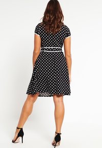 Svart klänning med prickar som har korta ärmar, en figurnära midja med ett bälte och en utsvängd kjol. Texturen verkar slät med en klassisk design.