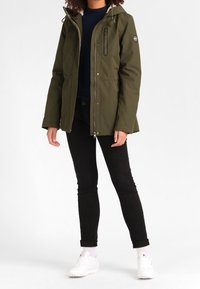 Veste imperméable vert olive avec fermeture éclair, capuche, poche poitrine et poignets élastiques, associée à un jean noir et des baskets blanches.
