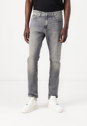 Mand iført falmede grå slim-fit jeans, sort skjorte og hvide sneakers, stående mod en ensfarvet hvid baggrund.