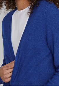 Persona con una blusa blanca y un cárdigan azul, sosteniendo el borde del cárdigan cerca del pecho, con el cabello rizado visible.