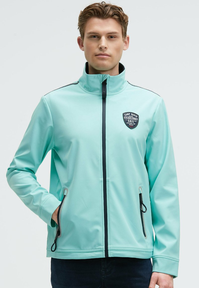 Giacca softshell verde menta con colletto alto, tasche con zip e un patch nero con logo sul petto. Texture liscia e design aderente.