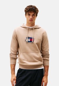 Joven con sudadera beige de Tommy Hilfiger con bolsillo frontal y pantalones azul marino, de pie contra un fondo claro y liso.