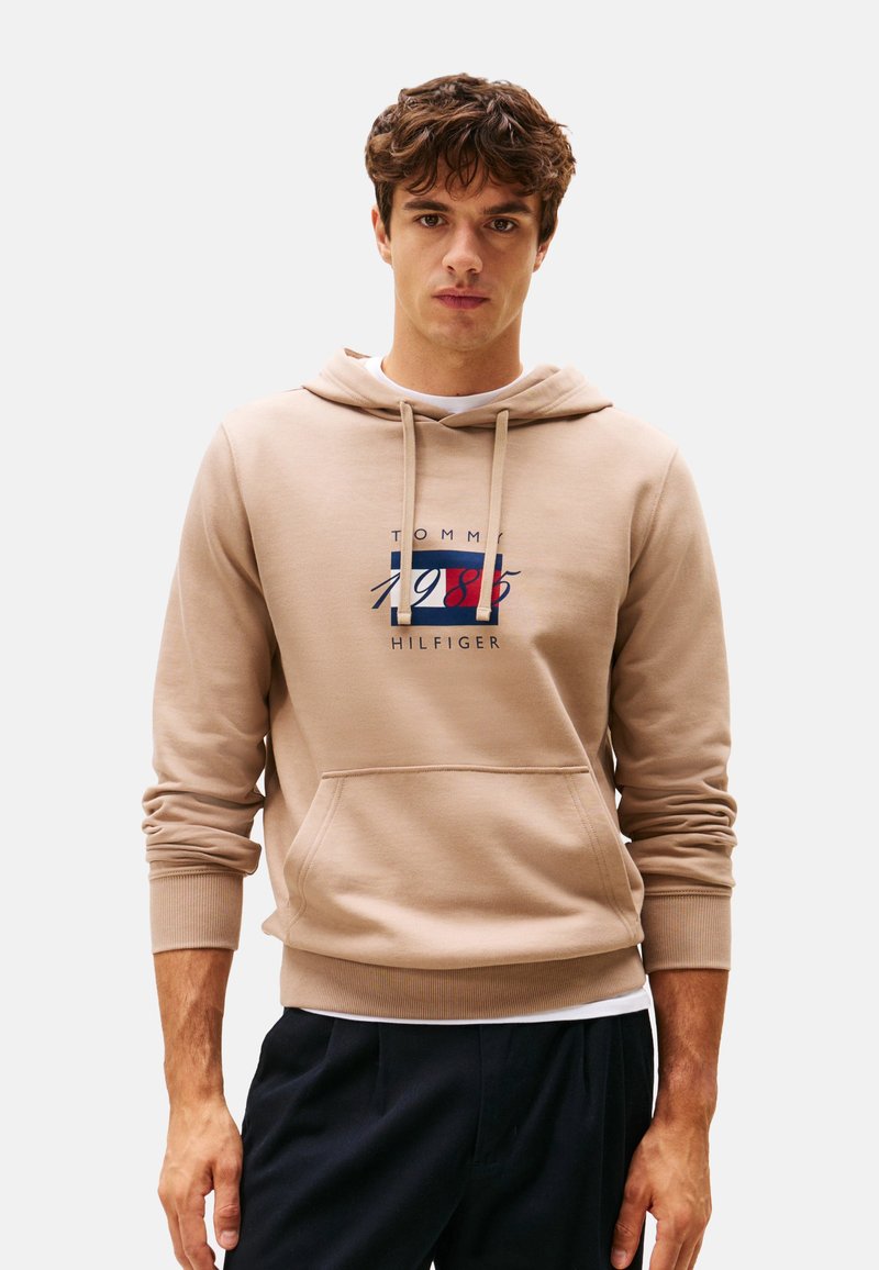 Joven con sudadera beige de Tommy Hilfiger con bolsillo frontal y pantalones azul marino, de pie contra un fondo claro y liso.