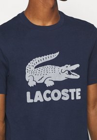 Osoba v námořnickém modrém tričku s velkým šedým logem krokodýla a textem "LACOSTE" vytištěným na přední straně.