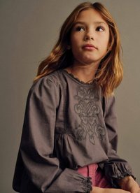 Blusa grigia con dettagli ricamati, maniche arricciate e scollatura a scallop, abbinata a pantaloni rosa. Tessuto morbido con una finitura testurizzata.