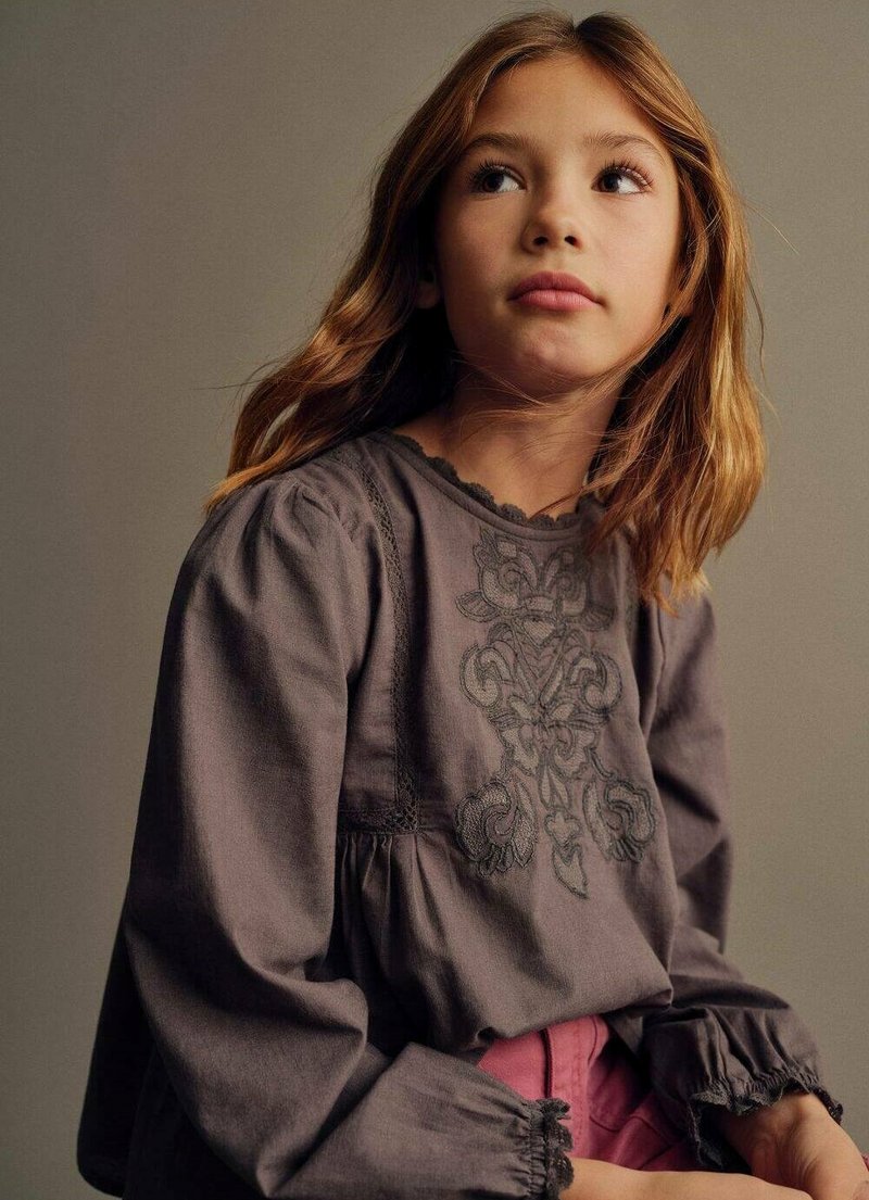 Blusa grigia con dettagli ricamati, maniche arricciate e scollatura a scallop, abbinata a pantaloni rosa. Tessuto morbido con una finitura testurizzata.