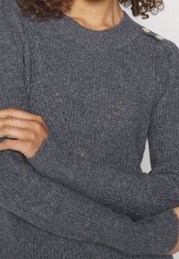 Pull gris en tricot avec un col rond, tissu texturé, accents de boutons sur l'épaule et motifs décoratifs ajourés.