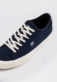 Sneaker in tela navy con lacci bianchi, suola in gomma testurizzata e un logo sul lato. Presenta un design a bassa altezza con tacco rinforzato.