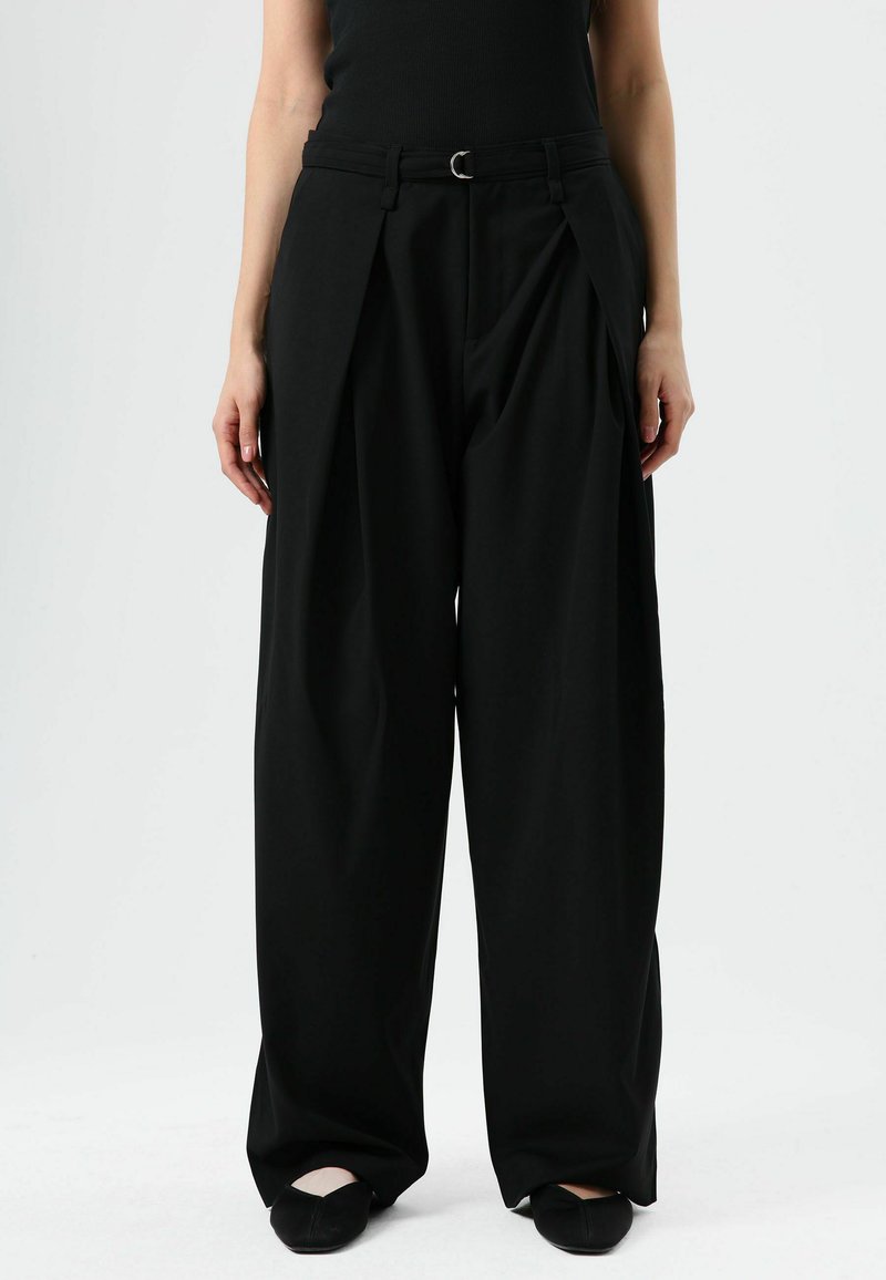 Pantalon noir à jambes larges avec une taille haute, présentant des plis à l'avant et une ceinture avec une boucle circulaire. Tissu lisse et texturé.