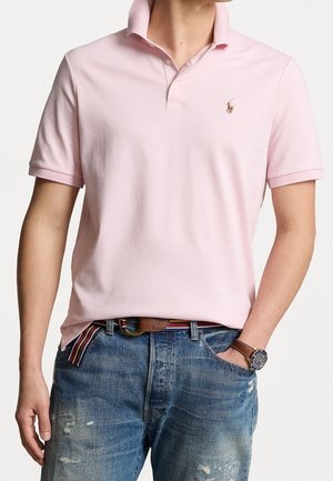 Mann trägt ein hellrosa Polohemd mit kurzen Ärmeln, blaue Jeans mit Used-Look, gestreiften Gürtel und eine Armbanduhr mit braunem Lederarmband.