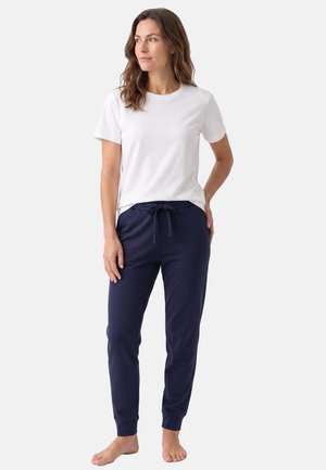 Femme debout pieds nus portant une chemise blanche à manches courtes et un pantalon de jogging bleu marine à cordon, sur un fond uni.