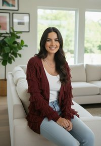 Bordowy dzianinowy kardigan z ozdobnymi frędzlami, noszony na białej bluzce, w połączeniu ze światło-niebieskimi dżinsami, siedzący na jasnej sofie.