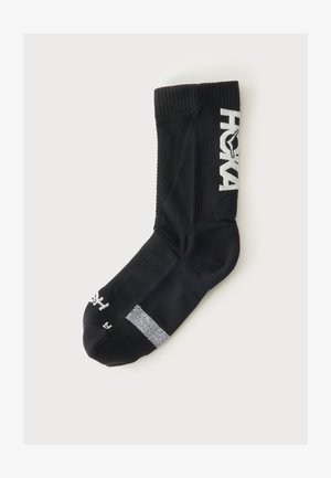 Chaussette noire style crew avec logo texte blanc sur le mollet et bande réfléchissante près des orteils, posée à plat sur un fond blanc.