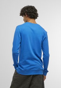 Camicia a maniche lunghe di un blu brillante, con collo rotondo e texture morbida, dal taglio rilassato. La vista da dietro mostra cuciture minime e design semplice.