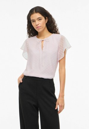 Femme aux cheveux bouclés portant un chemisier transparent rose clair à manches courtes avec un nœud papillon et un pantalon noir taille haute, debout devant un fond blanc.