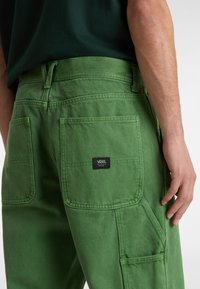 Green cargo pants with two large back pockets, visible stitching details, and a Vans logo tag on the right pocket. Textured cotton fabric.